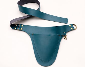 May include: Teal leather apron with a long strap and a pocket. The apron has brass rivets and a brass ring. The strap is gray on the underside. The apron is designed to be worn around the waist.