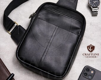 May include: Black leather crossbody bag with a zippered main compartment and an adjustable strap. The bag features a front pocket and a stitched vertical seam. A wristwatch and a smartphone are also visible.