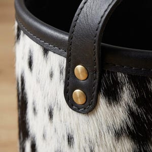 May include: A cylindrical container featuring a black and white spotted animal hide texture. The top edge is finished with a smooth black leather trim. A vertical black leather strap is secured to the side with two circular brass-toned metal rivets.