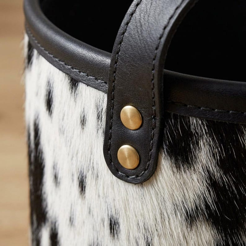 May include: A cylindrical container featuring a black and white spotted animal hide texture. The top edge is finished with a smooth black leather trim. A vertical black leather strap is secured to the side with two circular brass-toned metal rivets.
