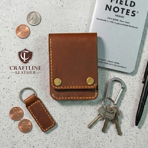 May include: A brown leather wallet with brass-colored snap closures, a matching keychain, and a Field Notes brand notebook. The wallet and keychain are stitched with yellow thread. US coins and keys are also present.