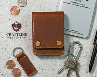 May include: A brown leather wallet with brass-colored snap closures, a matching keychain, and a Field Notes brand notebook. The wallet and keychain are stitched with yellow thread. US coins and keys are also present.