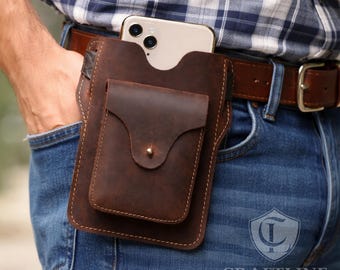 May include: A brown leather phone holster with a smaller front pocket, secured with a brass stud. The holster is attached to a brown leather belt, with a phone visible in the main compartment. The Craftline Leather logo is visible.