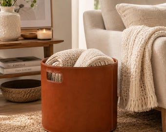 May include: A cylindrical storage bin made of smooth brown faux leather with integrated side handles. The bin features visible white stitching along the seams and contains a folded, cream-colored chunky knit textile inside the main compartment.