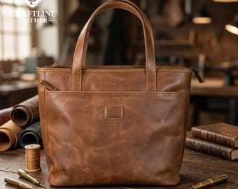 May include: A brown leather tote bag with two handles, a front pocket, and a zipper closure. The bag has a rectangular shape and a textured surface. The Craftline Leather logo is visible in the upper left corner. The bag is on a wooden surface.