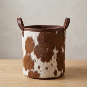 May include: A cylindrical storage basket featuring a brown and white cowhide pattern with a smooth brown leather rim and two matching leather handles attached with gold-toned metal rivets. The interior is lined with a dark fabric material.