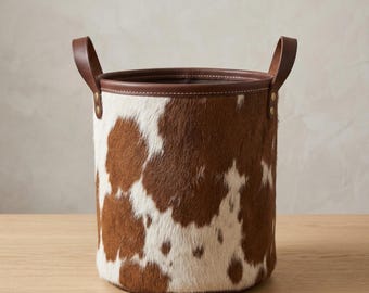 May include: A cylindrical storage basket featuring a brown and white cowhide pattern with a smooth brown leather rim and two matching leather handles attached with brass-colored metal rivets on opposite sides of the top edge.