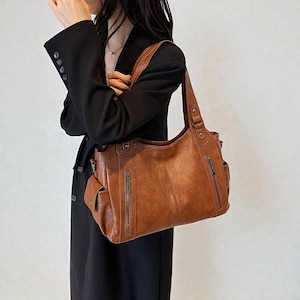 Könnte beinhalten: Eine braune Lederhandtasche mit mehreren Reißverschlusstaschen und einem Schulterriemen. Die Tasche hat ein strukturiertes Design mit einem leicht abgenutzten Aussehen. Die Tasche wird von einer Person getragen, die einen schwarzen Blazer trägt.