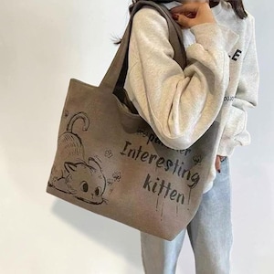 May include: A gray tote bag with a cartoon cat illustration and the text "Interesting kitten." The bag has a shoulder strap and is made of a canvas-like material. The cat illustration is black and the text is in a handwritten style.