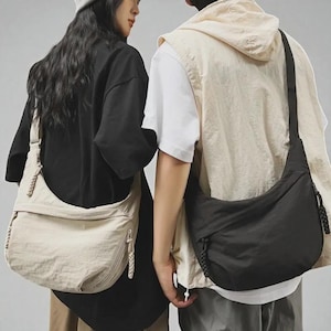 Puede incluir: Dos personas visten atuendos informales, cada una con un bolso cruzado. Un bolso es beige, el otro es negro. Ambos bolsos tienen forma curva, cremallera y detalle de cordón tejido en la correa.