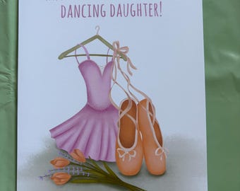 Op de afbeelding: Verjaardagskaart met een roze balletjurk, balletschoenen en tulpen. De kaart heeft de tekst "HAPPY BIRTHDAY TO A SUPER DANCING DAUGHTER!". De kaart ligt op een groene ondergrond.