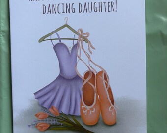 Op de afbeelding: Een verjaardagskaart met de tekst "HAPPY BIRTHDAY TO A SUPER DANCING DAUGHTER!". De kaart toont een paarse balletjurk op een hanger, een paar balletschoenen en een boeket oranje tulpen.