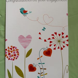 May include: Engagement card with a white background, featuring the text "Congratulations on your engagement." The card has a whimsical design with a blue bird, hearts, and floral elements in shades of red, pink, orange, and green.