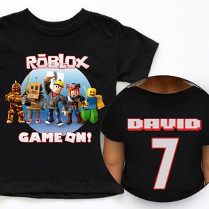 May include: Black t-shirt featuring a Roblox graphic with characters and the text "ROBLOX GAME ON!". The back of the shirt displays the name "DAVID" and the number "7".