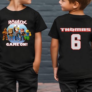 May include: Two children wearing black t-shirts. The front of one shirt features the word "ROBLOX" above a graphic of characters and the words "GAME ON!". The other shirt has the name "THOMAS" and the number "6".