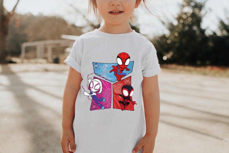 May include: A white t-shirt featuring a colorful graphic of three Spider-Man characters: Spider-Man, Spider-Gwen, and Miles Morales. The design is set against a backdrop of geometric shapes in blue, pink, and red. The shirt is worn by a young child.