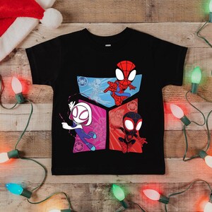 May include: Black t-shirt featuring Spider-Man characters. The shirt has a colorful graphic of Spider-Man, Spider-Gwen, and Miles Morales. A Santa hat and Christmas lights are in the background.