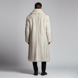 May include: A long, light-colored fur coat with a wide collar. The coat is worn over black pants and shoes. The image is taken from the back, showing the full length of the coat and the person wearing it.