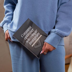 May include: A person holding a book with the title "Comment optimiser son compte Instagram" in white text on a black cover. The person is wearing a blue sweatshirt and matching pants. The background is a light gray.