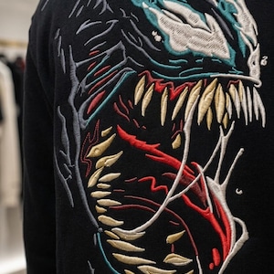 Könnte beinhalten: Schwarzer Kapuzenpullover mit detaillierter Stickerei eines Monstergesichts. Die Stickerei verwendet türkisfarbene, weiße, rote und gelbe Fäden, um das Bild von scharfen Zähnen und einem bedrohlichen Ausdruck zu erzeugen. Der Kapuzenpullover ist dunkel.