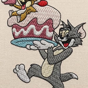 May include: Embroidered image of Tom and Jerry cartoon characters. Tom, a gray cat, carries a large cake on a blue plate. Jerry, a brown mouse, sits atop the cake, holding a cupcake. The cake has pink and red frosting.