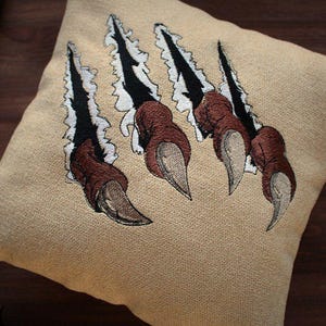 Pode incluir: Almofada quadrada bege com um desenho bordado de quatro marcas de garras castanhas e pretas rasgando o tecido. As garras têm pontas afiadas prateadas. A almofada é um artigo decorativo.