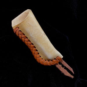 May include: A handmade leather finger protector in a light tan color, with a braided brown leather accent. The finger protector has two brown leather prongs extending from the end.