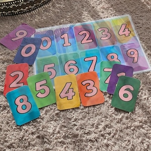May include: A colorful number matching game with a clear plastic overlay. The game includes a board with numbers 0-9 and individual cards with corresponding numbers. The cards are in various colors, including red, green, and yellow.