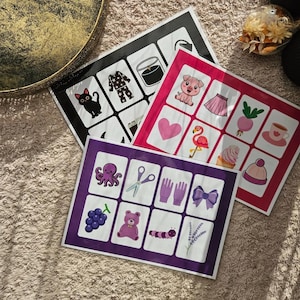 May include: Three colorful flashcards with various illustrations. One card is black with white images, another is pink with pink and red images, and the third is purple with purple and pink images. The cards are arranged on a beige surface.