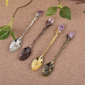 May include: Four decorative spoons with floral designs and amethyst crystal accents. The spoons have different metallic finishes: silver, bronze, gold, and antique gold. The spoons are arranged on a brown surface with green leaves in the background.