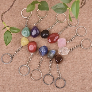 May include: A collection of keychains with various colorful gemstone pendants. The pendants are attached to silver-colored chains and key rings. Gemstones include green, purple, black, red, pink, yellow, blue, and clear stones. The background is a light brown fabric.