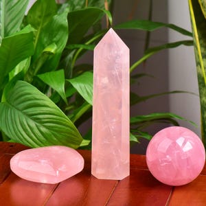May include: Three rose quartz crystals: a polished heart-shaped stone, a tall obelisk, and a sphere. The crystals are a soft pink color and sit on a wooden surface, with green foliage in the background.