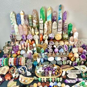 May include: A large collection of crystals and gemstones in various shapes and colors. The assortment includes polished spheres, towers, bracelets, and carved figurines. The crystals range from clear to vibrant hues of purple, green, and orange.