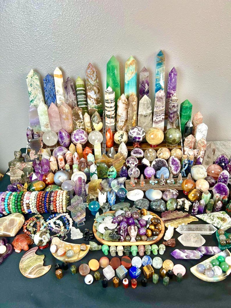 May include: A large collection of crystals and gemstones in various shapes and colors. The assortment includes polished spheres, towers, bracelets, and carved figurines. The crystals range from clear to vibrant hues of purple, green, and orange.