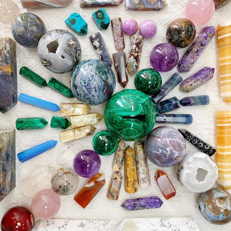 May include: A collection of various polished gemstones and crystals in different shapes and colors. The assortment includes spheres, points, and raw crystal formations in shades of green, purple, pink, and blue. The arrangement is on a white surface.