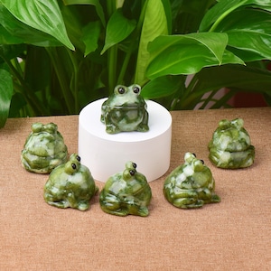 May include: A collection of green and white frog figurines. One frog sits atop a white cylinder, while five others are arranged around it. The frogs have detailed features and a polished finish, perfect for home decor.