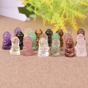 May include: A collection of small, carved Buddha figurines in various colors, including purple, clear, pink, green, and brown. The figurines are arranged on a tan surface, with a blurred background of green and yellow foliage.