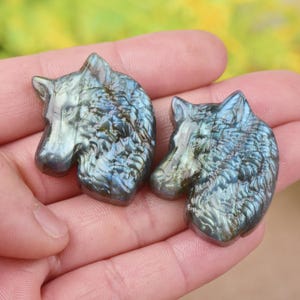 May include: Two labradorite wolf head carvings. The carvings are gray with iridescent blue and green flashes. The wolf heads are detailed with carved fur and facial features. The carvings are held in a person's hand.
