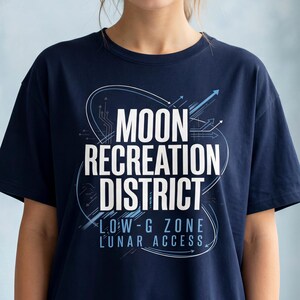 Puede incluir: Camiseta azul marino con el texto blanco "MOON RECREATION DISTRICT" y el texto adicional "LOW-G ZONE LUNAR ACCESS". La camiseta tiene cuello redondo y mangas cortas.