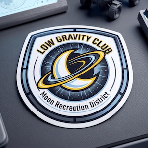 May include: A sticker with a shield design featuring the text "LOW GRAVITY CLUB" and "Moon Recreation District". The sticker has a crescent moon and ring design in yellow and blue against a starry background.
