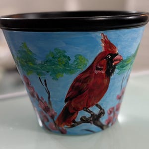 May include: A hand-painted ceramic flower pot with a black rim and a light blue background. The pot features a vibrant red cardinal perched on a branch, with green foliage and pink blossoms. The pot is approximately 13 cm tall.