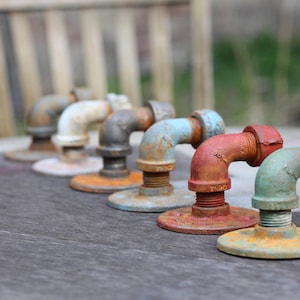 May include: A row of seven rusty metal pipe fittings in various colors, including white, gray, blue, red, and green. The fittings are arranged in a line on a wooden surface.