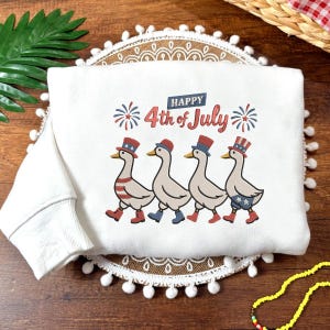 May include: White sweatshirt with a patriotic design for the 4th of July. The graphic features four geese wearing hats and boots in red, white, and blue. The text "Happy 4th of July" is printed above the geese.