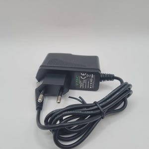 Könnte beinhalten: Ein schwarzes V-TAC-Netzteil mit einem zweipoligen Stecker und einem schwarzen Kabel. Der Adapter trägt das V-TAC-Logo und andere aufgedruckte Texte. Das Kabel ist aufgerollt und liegt auf einer weißen Oberfläche.