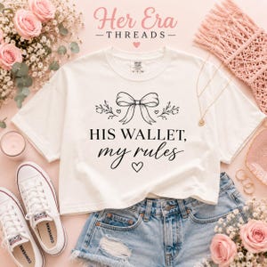 May include: A white crop top with the text "HIS WALLET, my rules" in black script. The top features a bow and floral design. The image includes denim shorts, white Converse sneakers, and floral arrangements.