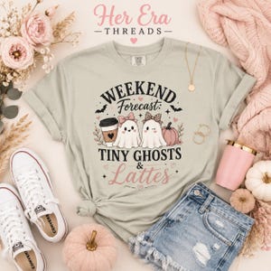 May include: A light sage green t-shirt featuring a graphic print with the text "WEEKEND Forecast: TINY GHOSTS & Lattes". The design includes two cartoon ghosts, a coffee cup, a pumpkin, bats, and small hearts in black, pink, and brown.