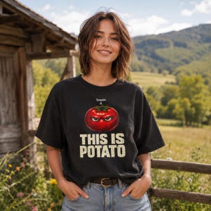 May include: A black short-sleeve t-shirt featuring a graphic of a red, angry-looking tomato with the word "Tomato" and an arrow pointing to it above, and the text "THIS IS POTATO" printed in large, distressed white capital letters below.