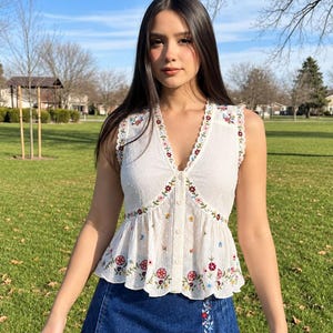 May include: A white sleeveless top with a V-neckline, button-down front, and colourful floral embroidery, paired with a dark blue denim mini skirt featuring a vertical floral embroidered panel on the side and a pleated hemline design.