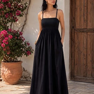 May include: A black maxi dress with thin straps and a square neckline. The dress has a fitted bodice and a flowing skirt with pockets. The model is wearing gold hoop earrings and sandals. The background includes a wooden door, a white wall, and a potted plant with pink flowers.