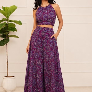 May include: A two-piece set consisting of a sleeveless halter-neck crop top and matching high-waisted wide-leg trousers. Both pieces feature a vibrant purple fabric with an intricate, repeating geometric and floral pattern in blue and gold tones.